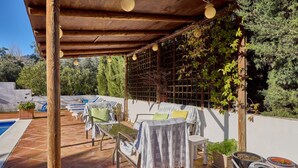 Zona d'estar a l'aire lliure coberta, amb piscina, una pèrgola de fusta i plantes en testos.