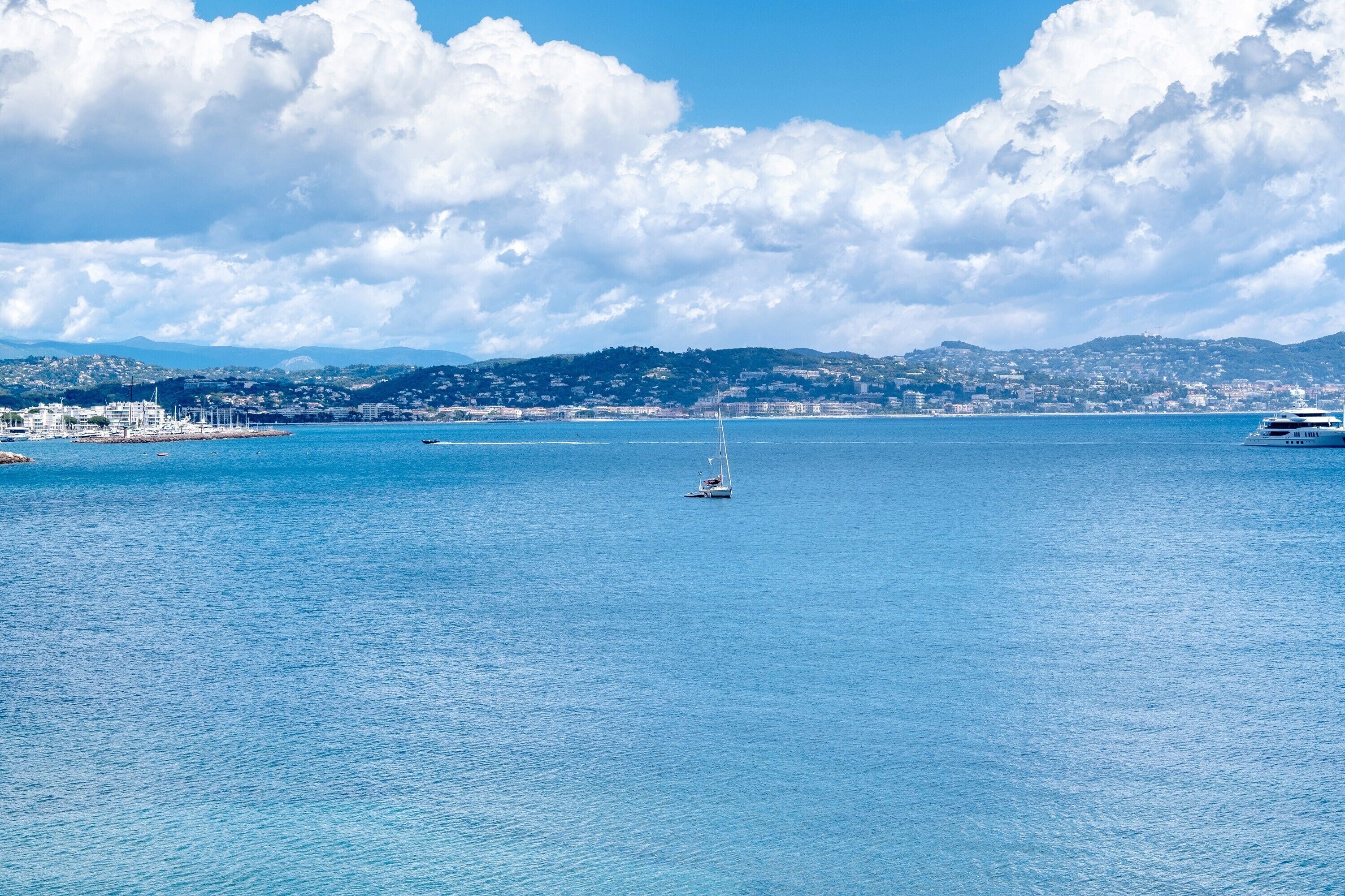 A coastal cityscape with a clear sky, calm sea, and a sailboat.