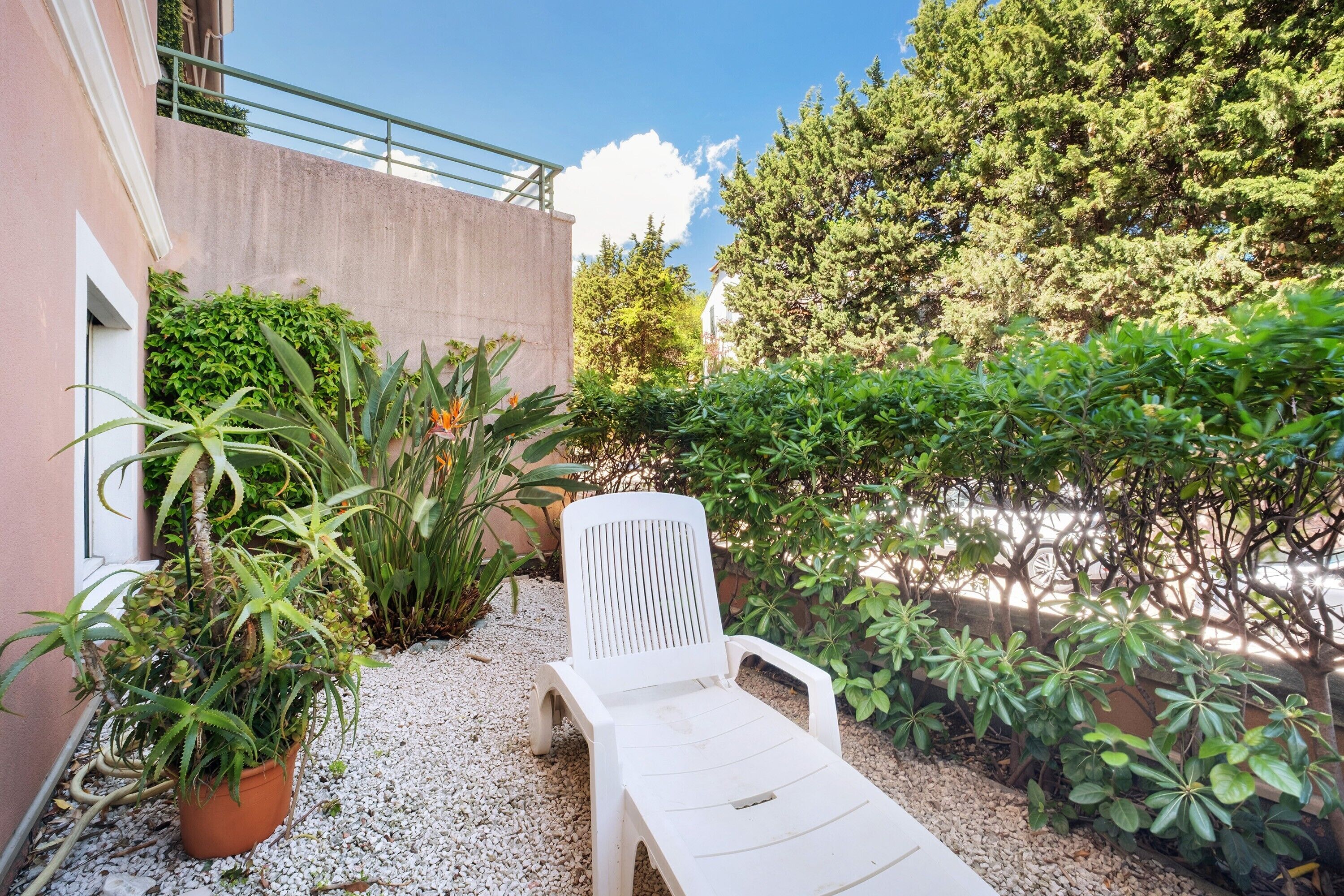 Una silla de lounge blanca en un jardín con un sendero de grava, rodeada de diversas plantas y un edificio con balcón.