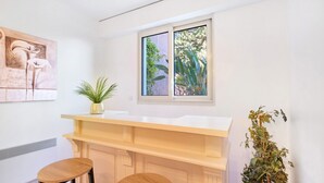 Eine moderne Küche mit weißem Arbeitsplatz, zwei Holz-Barhocker, einem Fenster mit Blick auf Grünflächen und einer Wandpflanze.