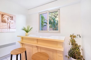 Cucina moderna con un piano di lavoro bianco, due sgabelli in legno, una finestra con vista sul verde e una pianta sospesa al muro.