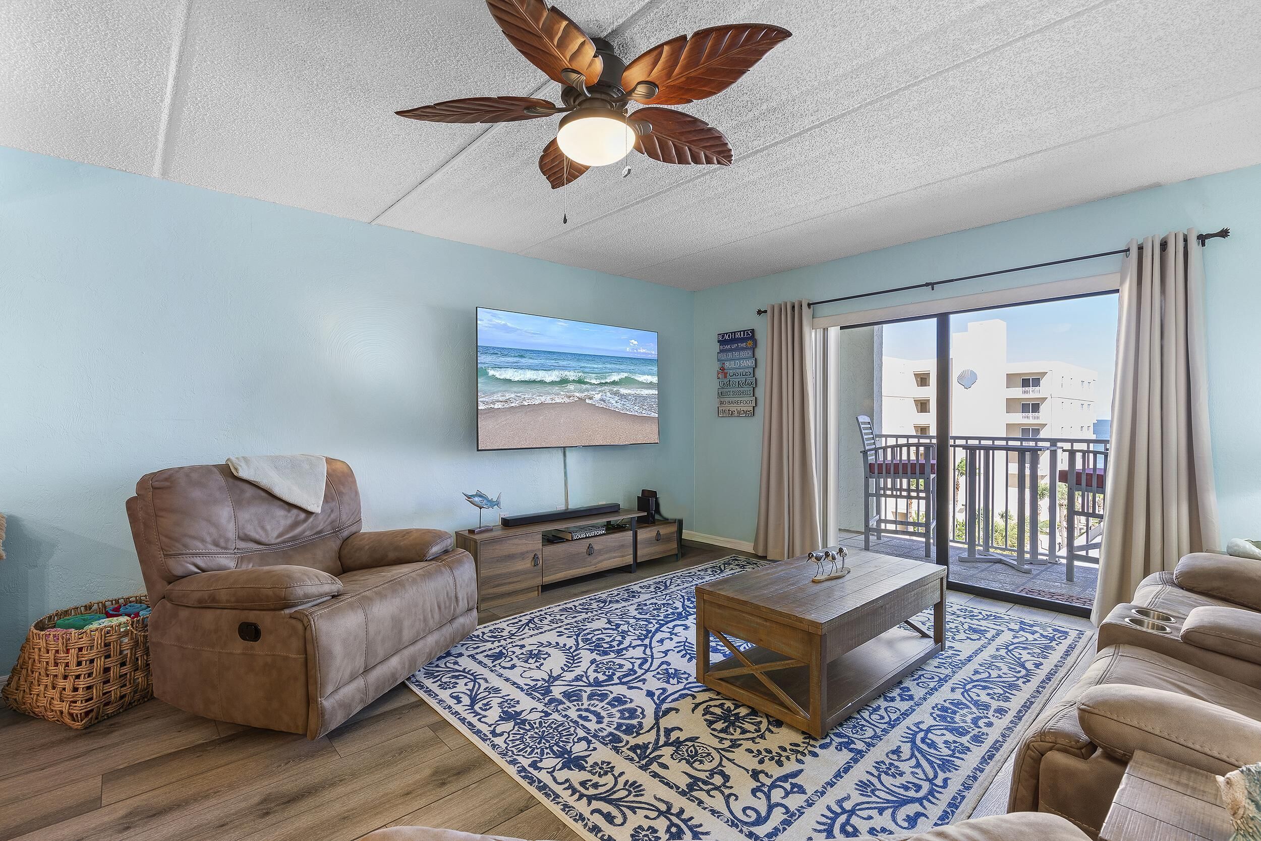 Ein Wohnzimmer mit Deckenventilator, großem Fernseher, einer Couch, einem Couchtisch und Blick auf den Strand durch eine Glastür.