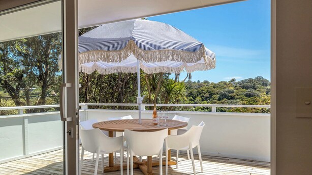 A wooden deck with a round table and white chairs, an umbrella, and a view of a lush landscape.