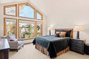 A bedroom with a large bed, two bedside tables with lamps, a chair, and a view of a building through large windows. - Private Hot Tub & Garage, Near Main St/Peak 9 (Breckenridge)
