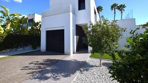 A modern two-story house with a garage, surrounded by a landscaped garden with palm trees and a small tree in the foreground. - Villa Levante, Modern Costa Blanca villa with pool and sunny terrace (San Fulgencio)