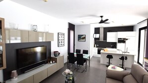 A modern living room with a flat-screen TV, a glass coffee table, and a sofa. There is a dining area with a table and chairs, and a kitchen with white cabinets and a black countertop. - Villa Levante, Modern Costa Blanca villa with pool and sunny terrace (San Fulgencio)