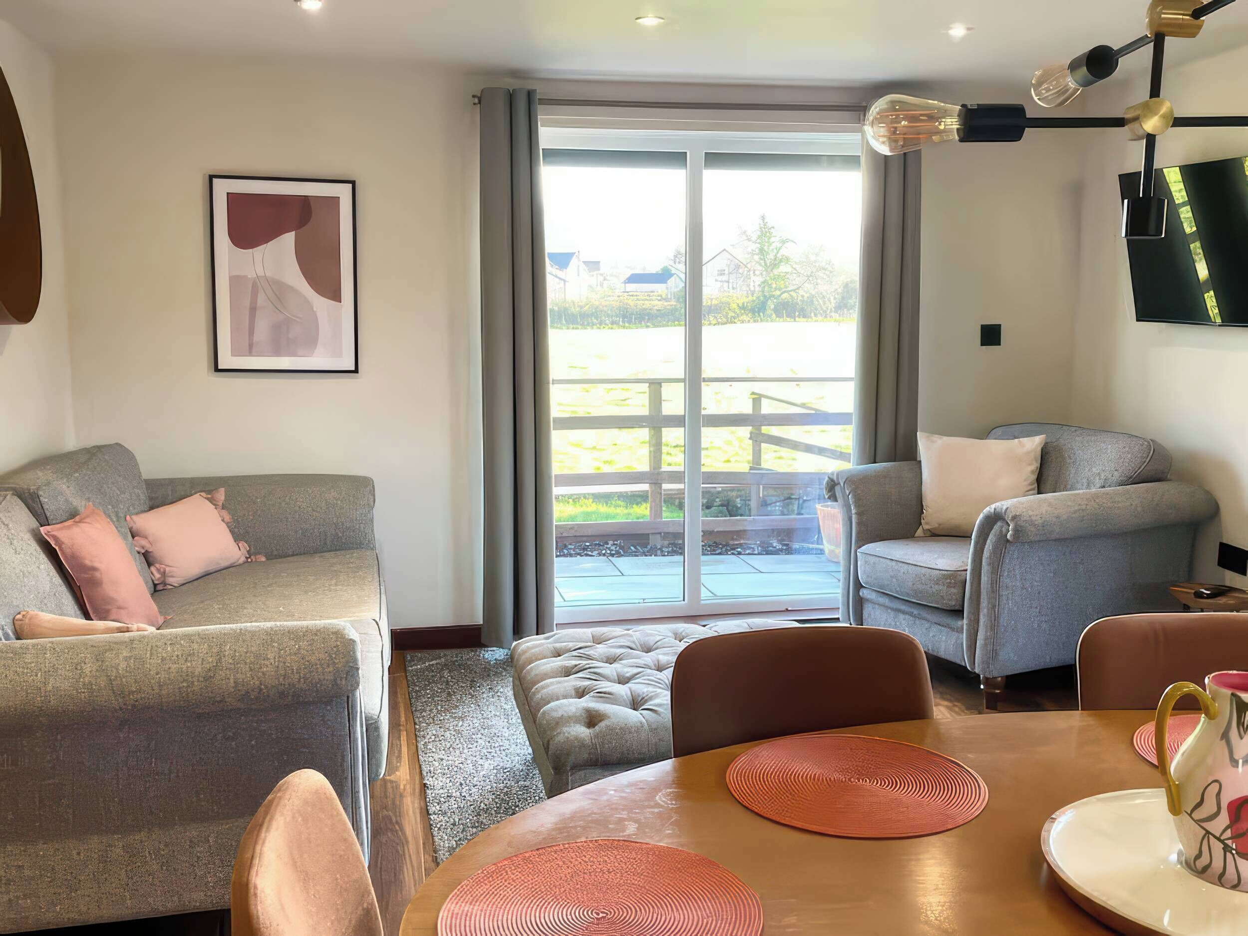 Una sala moderna con un sofá gris, un sillón azul y una mesa de comedor de madera con individuales. Se ve un ventanal con vistas a un jardín.