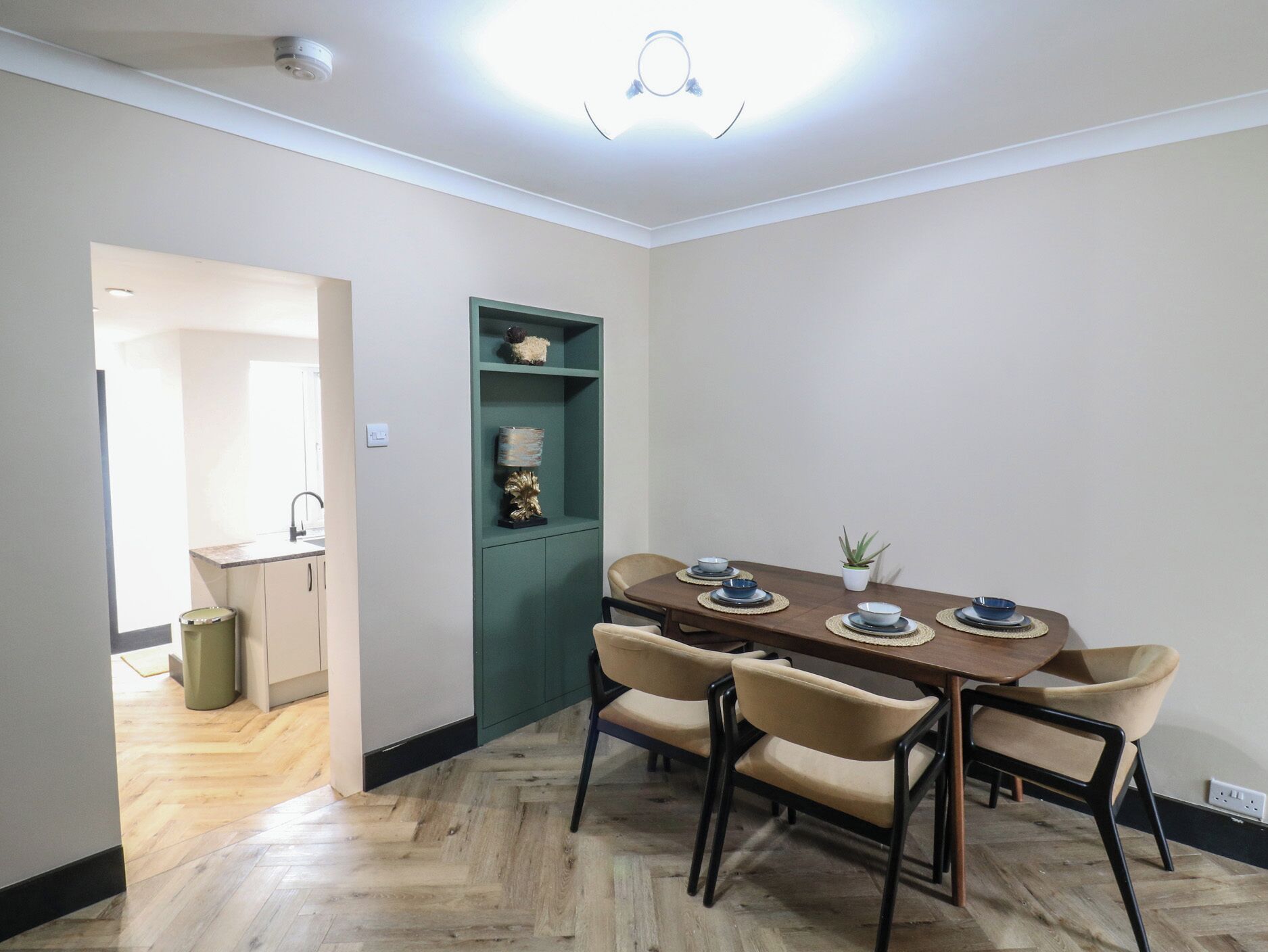 Un coin repas avec une table en bois et des chaises de couleur beige, un meuble vert agrémenté d’objets décoratifs et une petite plante en pot.