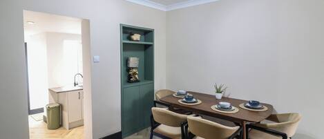 Un coin repas avec une table en bois et des chaises de couleur beige, un meuble vert agrémenté d’objets décoratifs et une petite plante en pot.