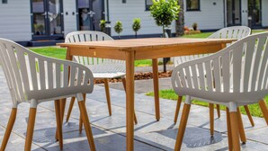 A patio with a wooden table and white plastic chairs, a house in the background, and a small tree in the center. - Silver Birch Cottage (Llanrwst)