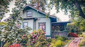 Una casa de dos pisos con base de piedra y un jardín con diversas plantas y flores.