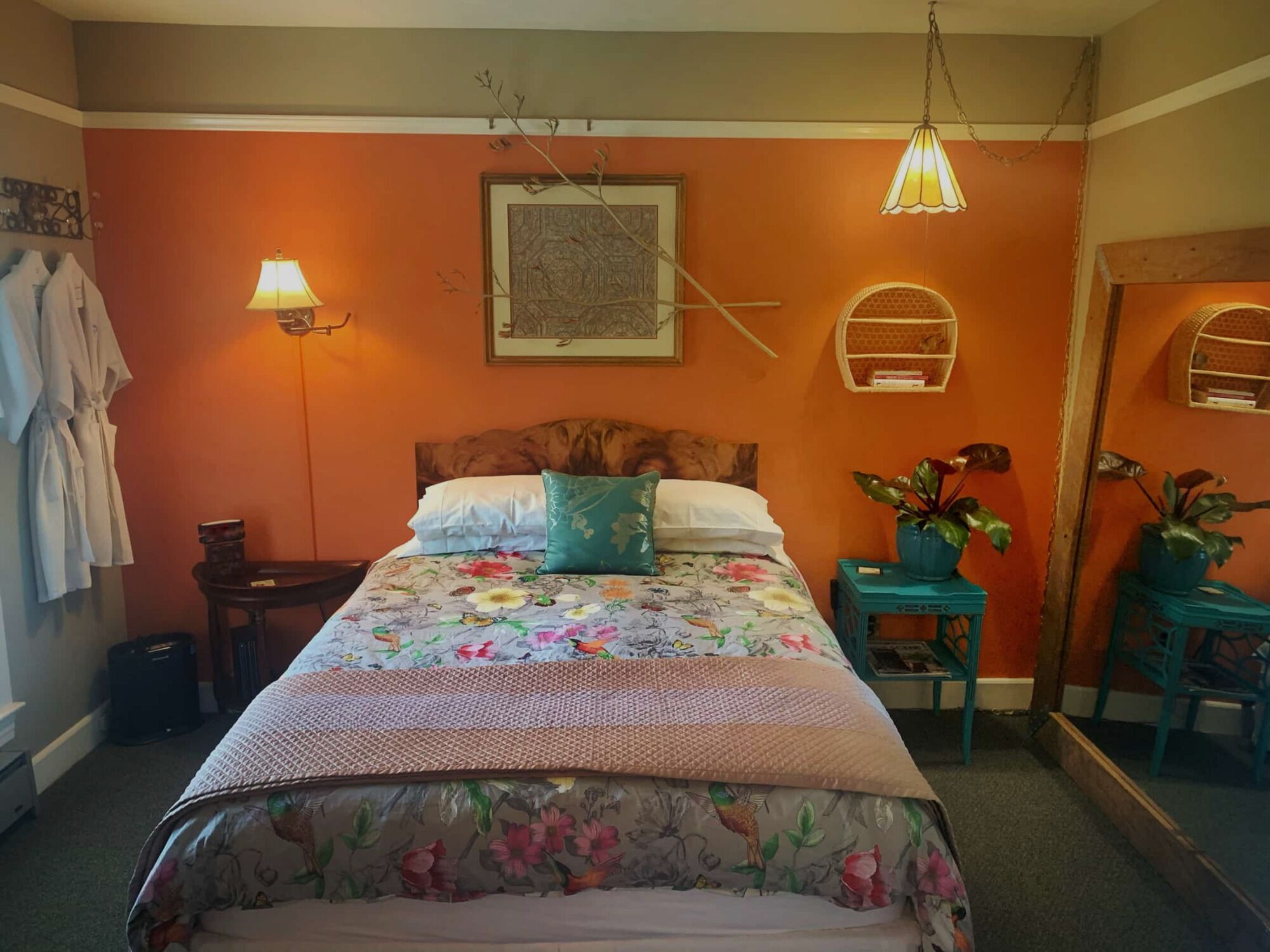 Une chambre avec un mur orange, une tête de lit en bois, une couvre-lit à motifs floraux, une table de chevet avec une lampe et un miroir.
