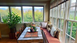Un coin repas avec une table en bois, des chaises et un banc agrémenté de coussins. Une grande fenêtre offre une vue sur les arbres et les montagnes.