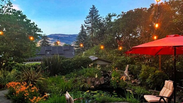 Une terrasse avec un sol en pierre, deux chaises, un parasol rouge et un bassin entouré de végétation.