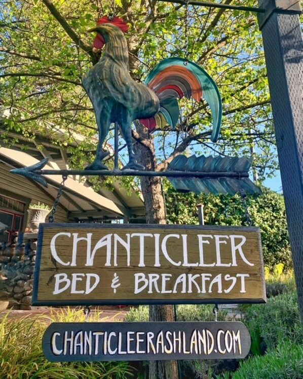 Puinen kyltti, jossa on kuvattuna kukko ja sateenkaari, ja jossa mainostetaan majataloa nimeltä ’Chanticleer Bed & Breakfast’ sekä ilmoitetaan sen verkkosivuston osoite.