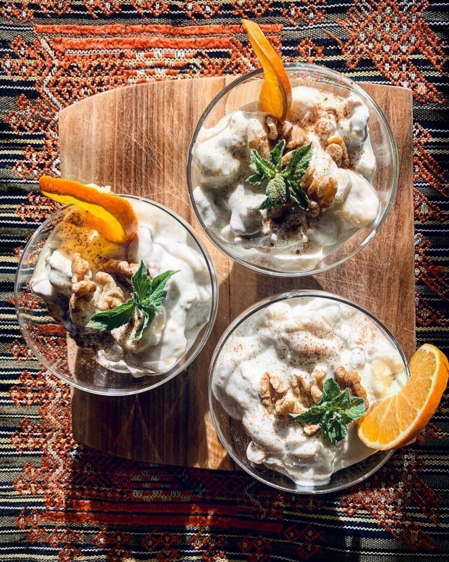 Three bowls of a creamy dessert topped with walnuts, orange slices, and mint leaves, placed on a wooden board.