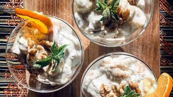 Three bowls of a creamy dessert topped with walnuts, orange slices, and mint leaves, placed on a wooden board.