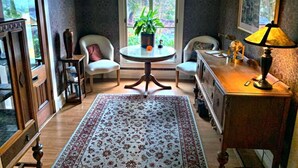 Ein gemütliches Zimmer mit einem runden Tisch, zwei Stühlen, einem Blumentopf, einer Lampe und einem großen Fenster mit Blick auf Grünflächen.