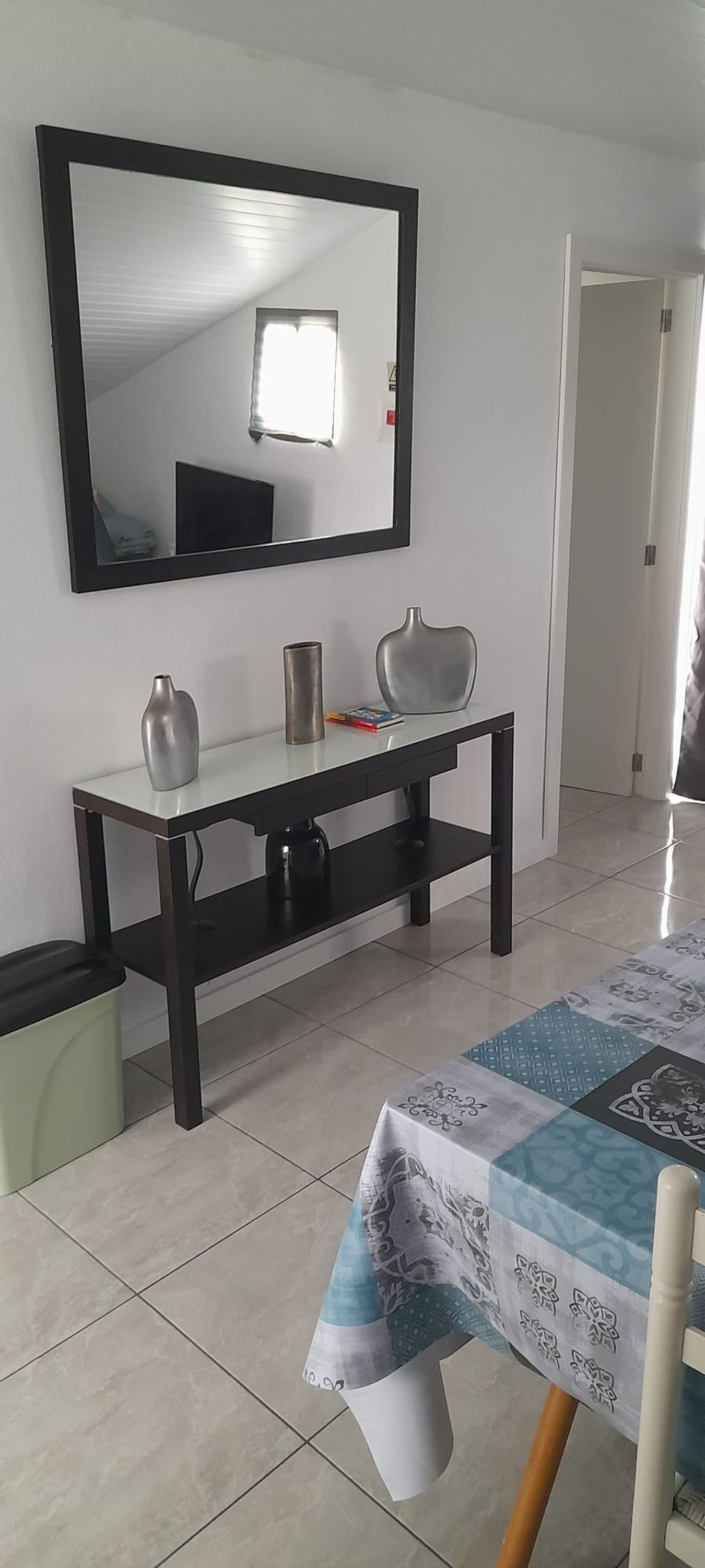 Ein Zimmer mit weiß gefliestem Boden, einem spiegeln schwarzen Rahmen, einem weißen Tisch mit gemustertem Tischläufer und einer schwarzen Konsole mit dekorativen Gegenständen.