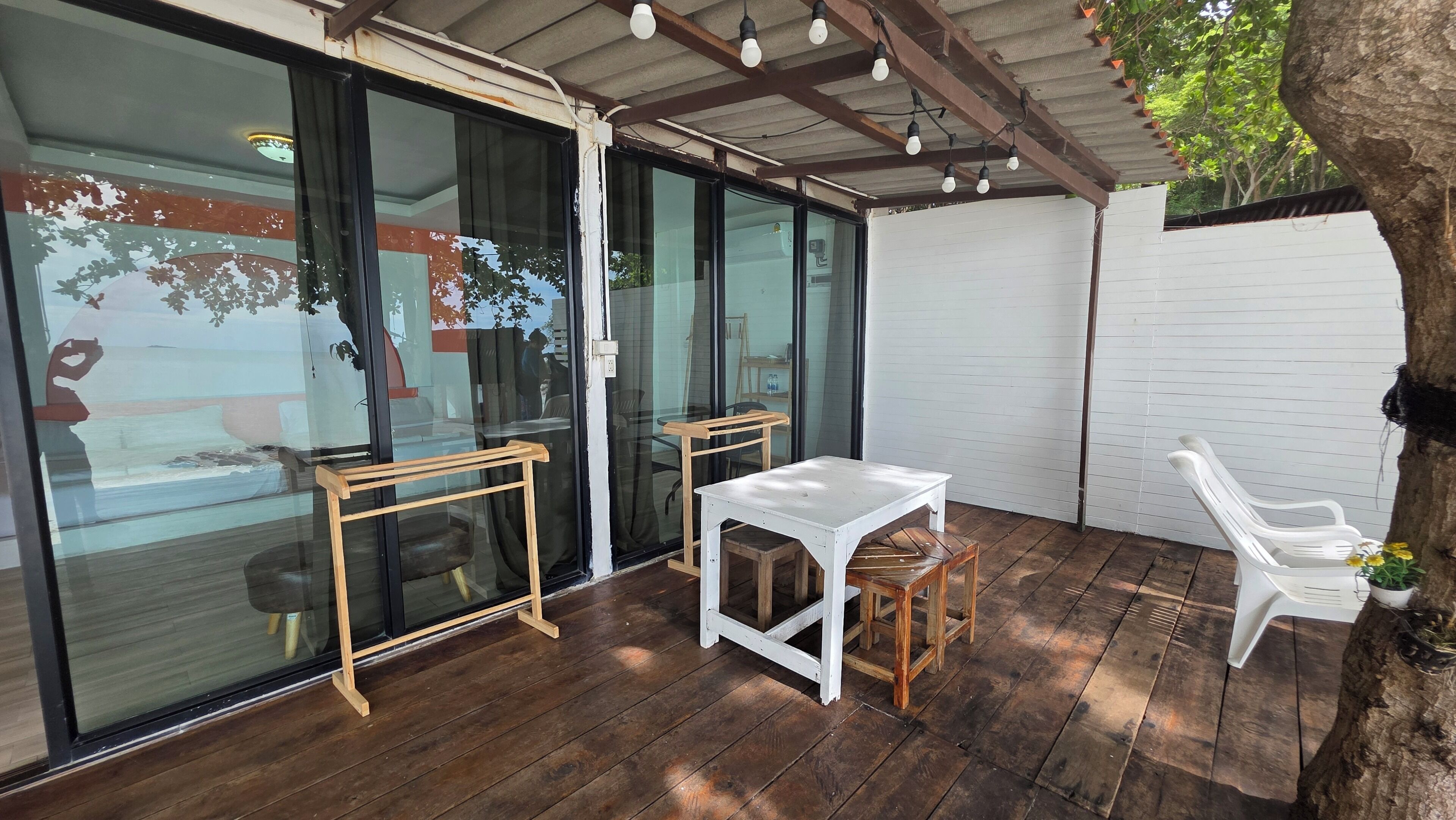 A covered patio with wooden flooring, a white table, two wooden chairs, and a white plastic chair.