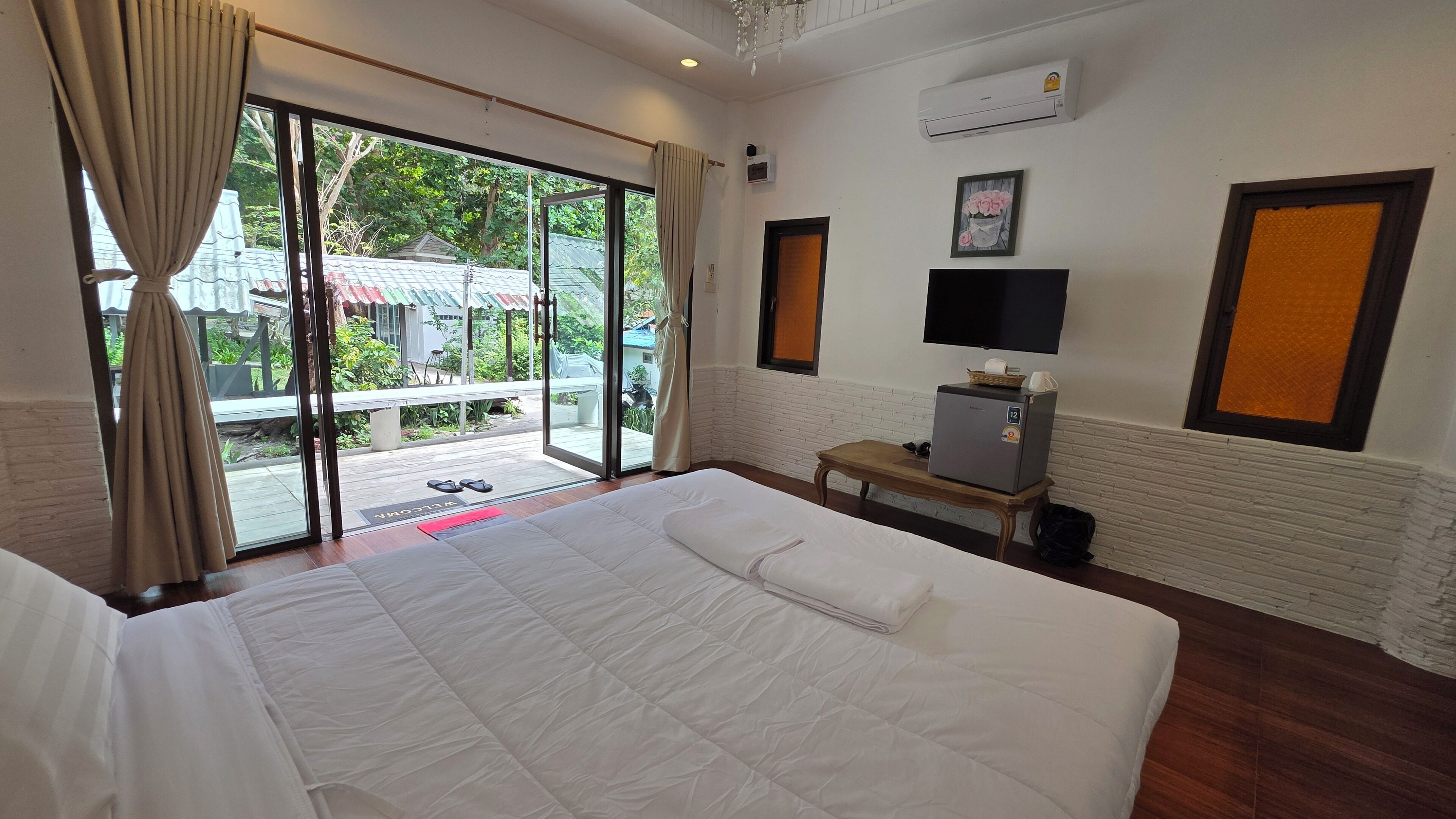 A hotel room with a large bed, a small desk with a refrigerator, a television, and a sliding glass door leading to an outdoor area.