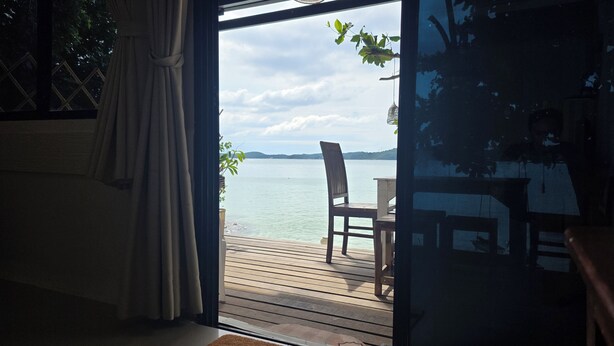 A wooden deck with a chair facing a calm body of water, a view of the sky, and a curtain partially drawn. - Samed Big Cape Resort (Rayong)