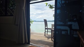 A wooden deck with a chair facing a calm body of water, a view of the sky, and a curtain partially drawn. - Samed Big Cape Resort (Rayong)