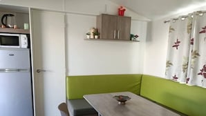 Eine kompakte Küchenzeile mit Mikrowelle und Kühlschrank, eine grüne Polsterbank mit Tisch sowie ein Fenster mit floralem Vorhang.