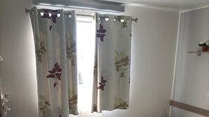 Ein Schlafzimmer mit einem Bett, einem Fenster mit floralem Vorhang, einem Nachttisch mit Lampe und einem kleinen Regal mit Pflanze.