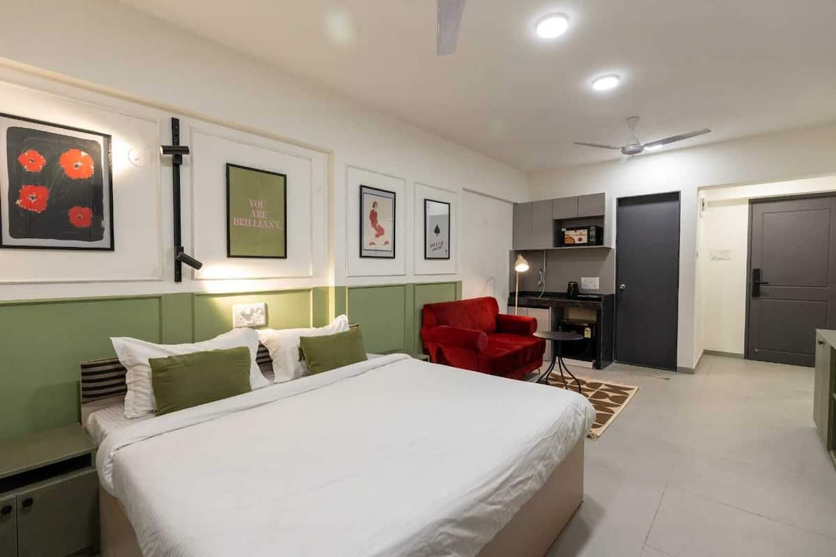 A hotel room with a bed, a red armchair, a desk, and a kitchenette.