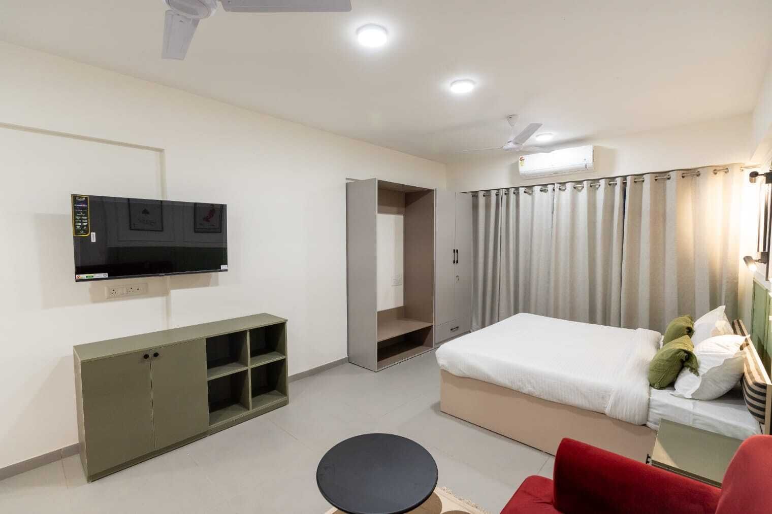 A hotel room with a bed, a TV mounted on the wall, a wardrobe, a desk with a chair, and a red armchair.