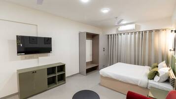 A hotel room with a bed, a TV mounted on the wall, a wardrobe, a desk with a chair, and a red armchair.