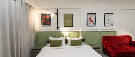 Une chambre d’hôtel avec un lit, une tête de lit verte, un fauteuil rouge et des tableaux encadrés accrochés au mur.