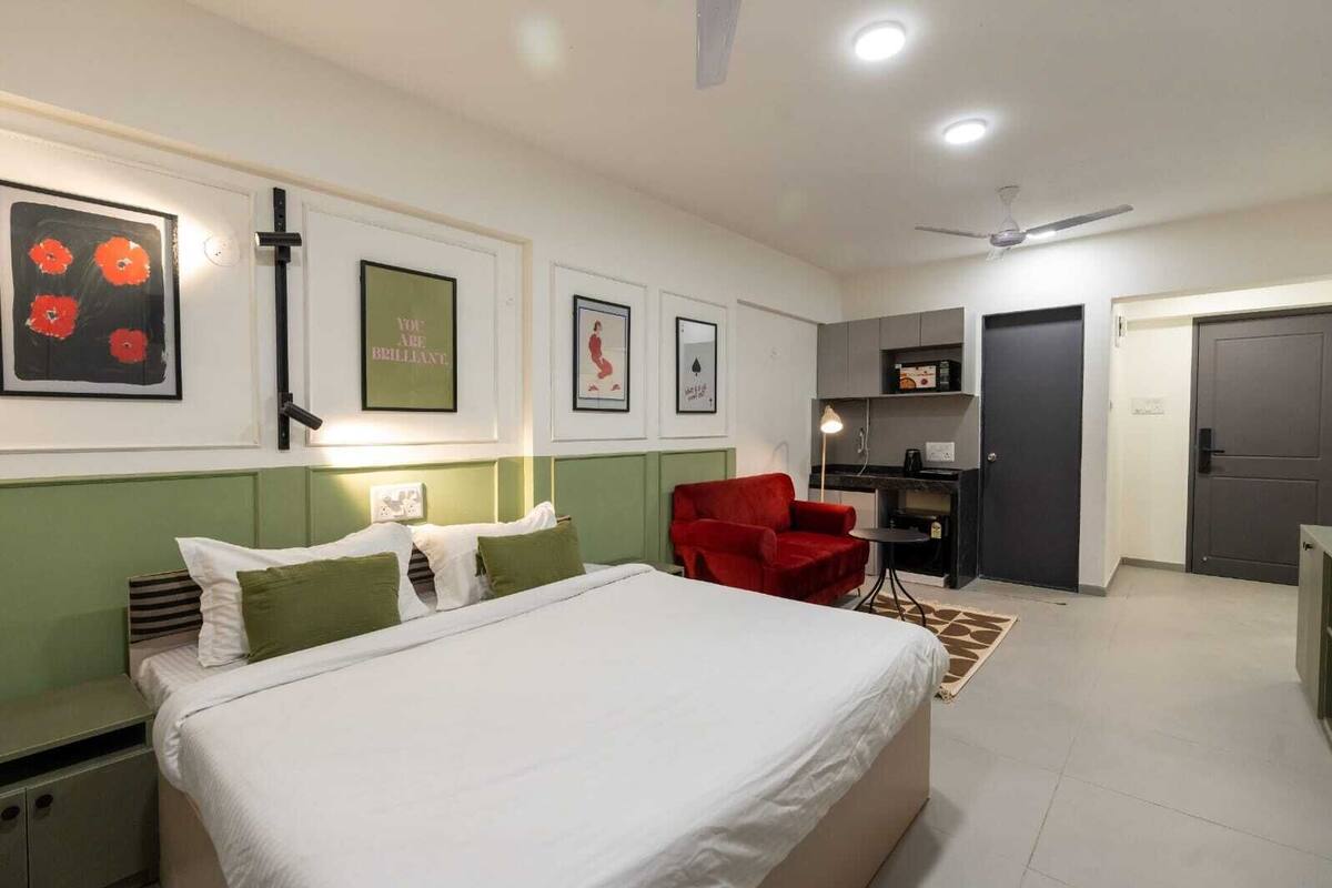 A hotel room with a bed, a red armchair, a desk, and a kitchenette.