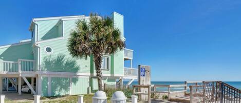 A beachfront property with a multi-level structure, a palm tree, and a wooden boardwalk leading to the beach.