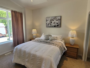 A bedroom with a large bed, two bedside lamps, a window with curtains, and a wall-mounted floral artwork. - Lakefront Winter Rental - Perfect for Snowbirds (Osoyoos)
