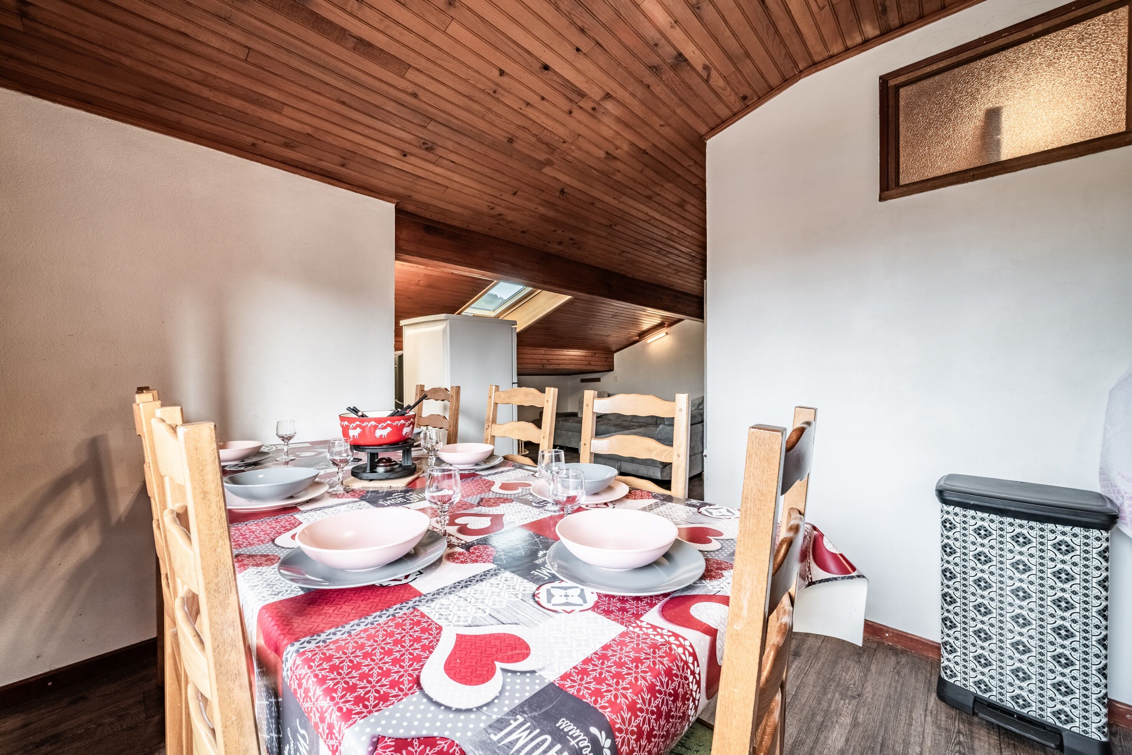 Een eethoek met een tafel voor vier personen, houten stoelen, een tafelkleed met dessin en een prullenbak.