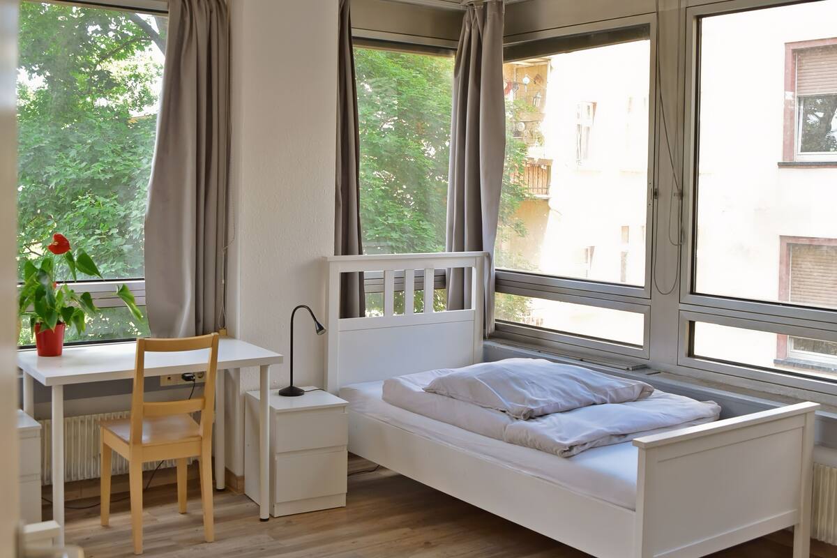 Un dormitorio con una cama blanca, un escritorio con silla, una mesita de noche y una planta en maceta.