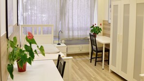 Una habitación pequeña y bien iluminada con una cama, un escritorio y una silla. Se ve una ventana con cortinas translúcidas, un radiador y una planta en maceta sobre el escritorio.