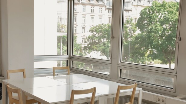 Un comedor con una mesa blanca y sillas de madera, ventanas amplias con vistas a edificios y árboles, y piso de baldosas.