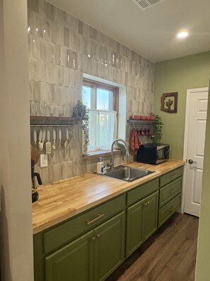 Una cocina con encimera de madera, armarios verdes, un lavamanos y una ventana con cortinas.