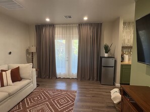 Una sala de estar con un sofá, un tapete estampado, una zona de cocina con un refrigerador y una ventana con cortinas.