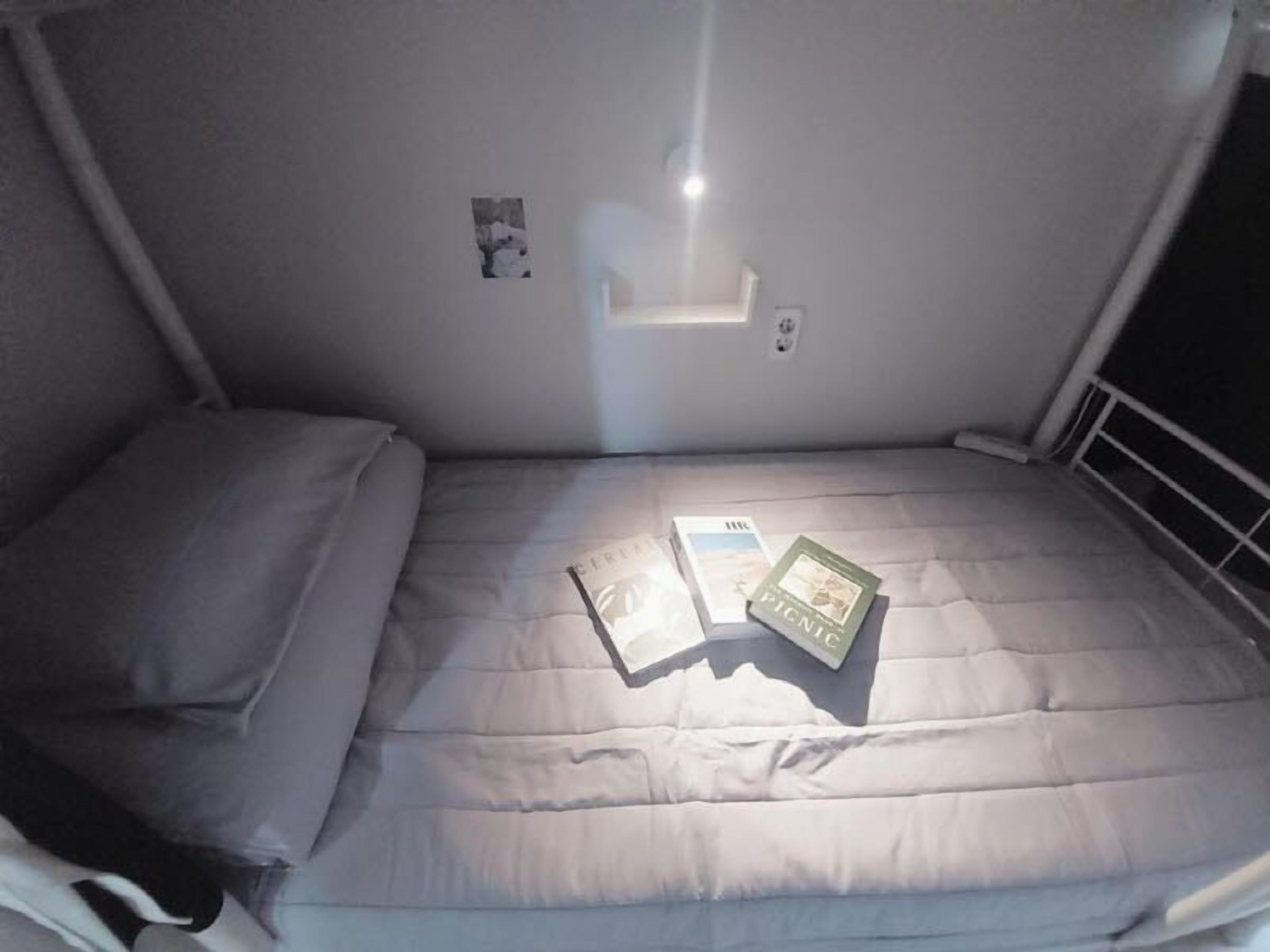 Una cama individual con un colchón gris y una almohada, una mesita de noche pequeña con un libro y una caja, y un aplique de pared.