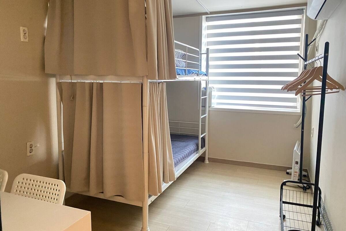 Asuntolan huone, jossa on kerrossängyt, ikkuna, jossa on sälekaihtimet, työpöytä ja tuoli.