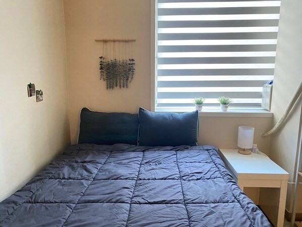 A bedroom with a bed, a window with blinds, a small desk with a lamp, and two potted plants on the windowsill. - Funwave guesthouse in Haeundae (Busan)