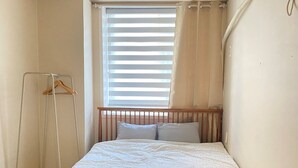 A small, neatly made bed with a wooden headboard in a corner room with a window and a clothes drying rack.