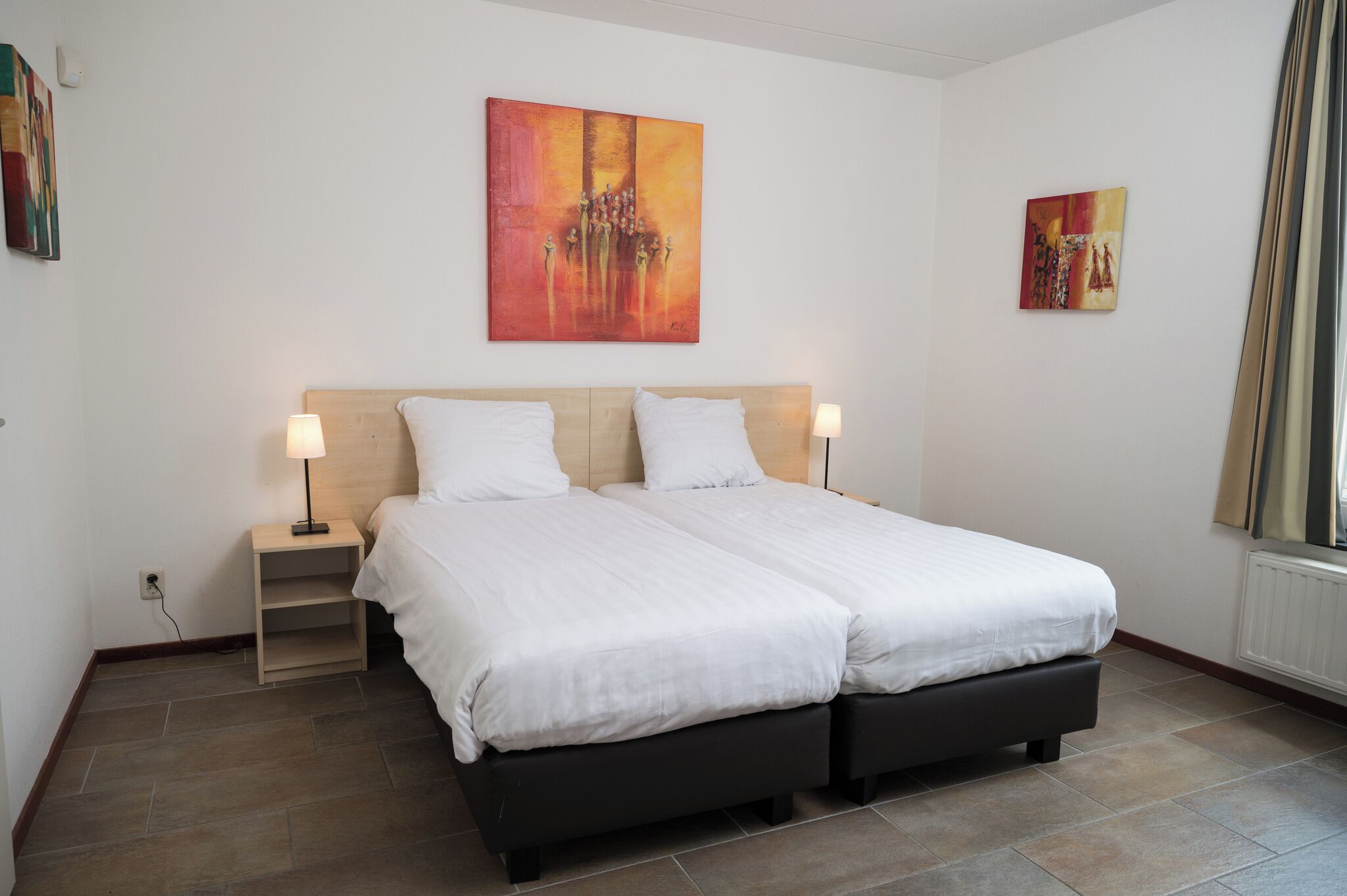 A double bed with two white pillows and a wooden headboard, flanked by nightstands with lamps, in a room with tiled flooring and two abstract paintings on the walls.