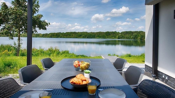 An outdoor dining area with a table set for two, overlooking a calm lake and surrounded by greenery. - Resort Boschmolenplas (Heel)