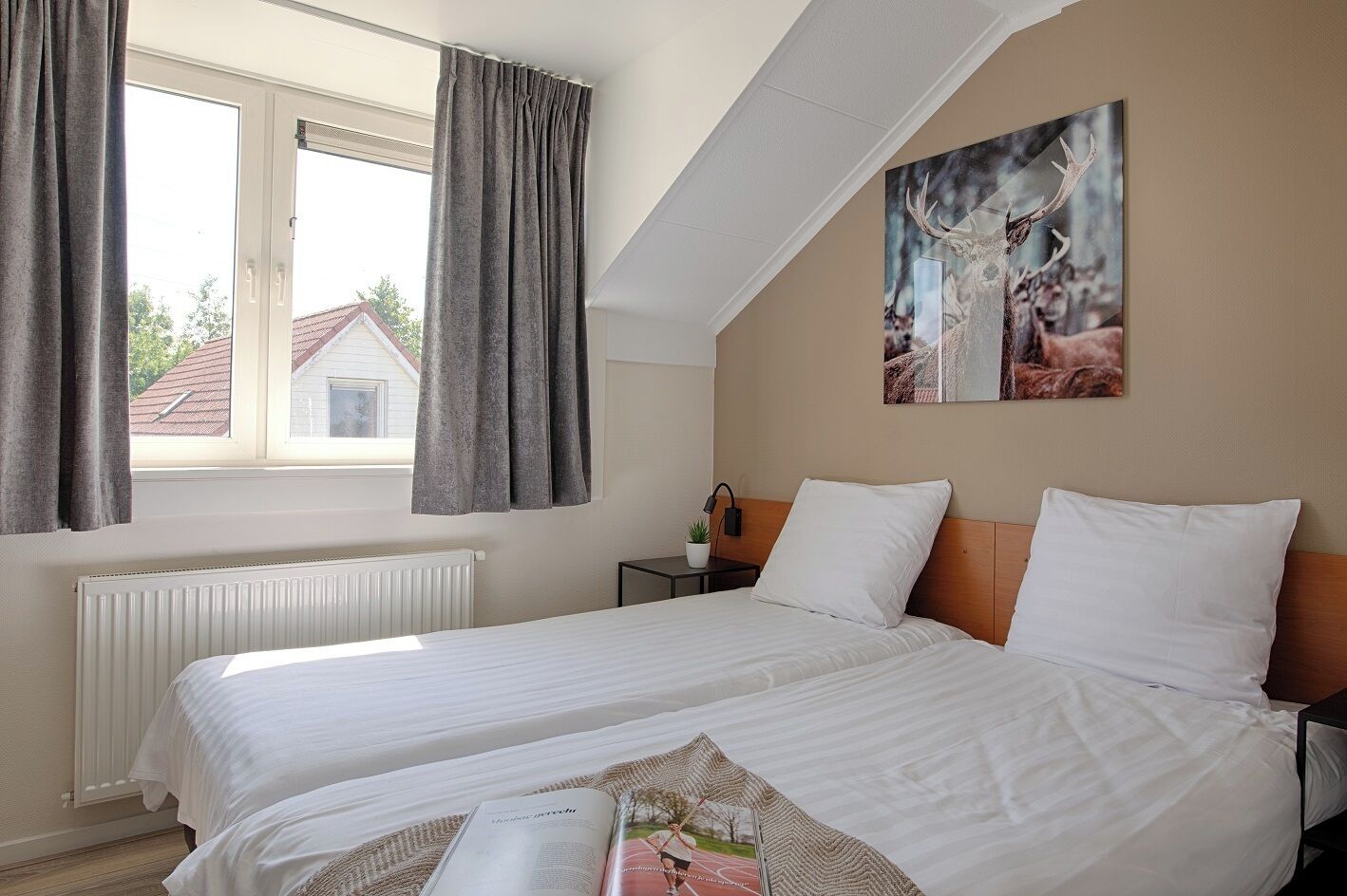 Een hotelkamer met een groot bed, een raam met gordijnen, een radiator, een nachtkastje met een lamp en een schilderij aan de muur.