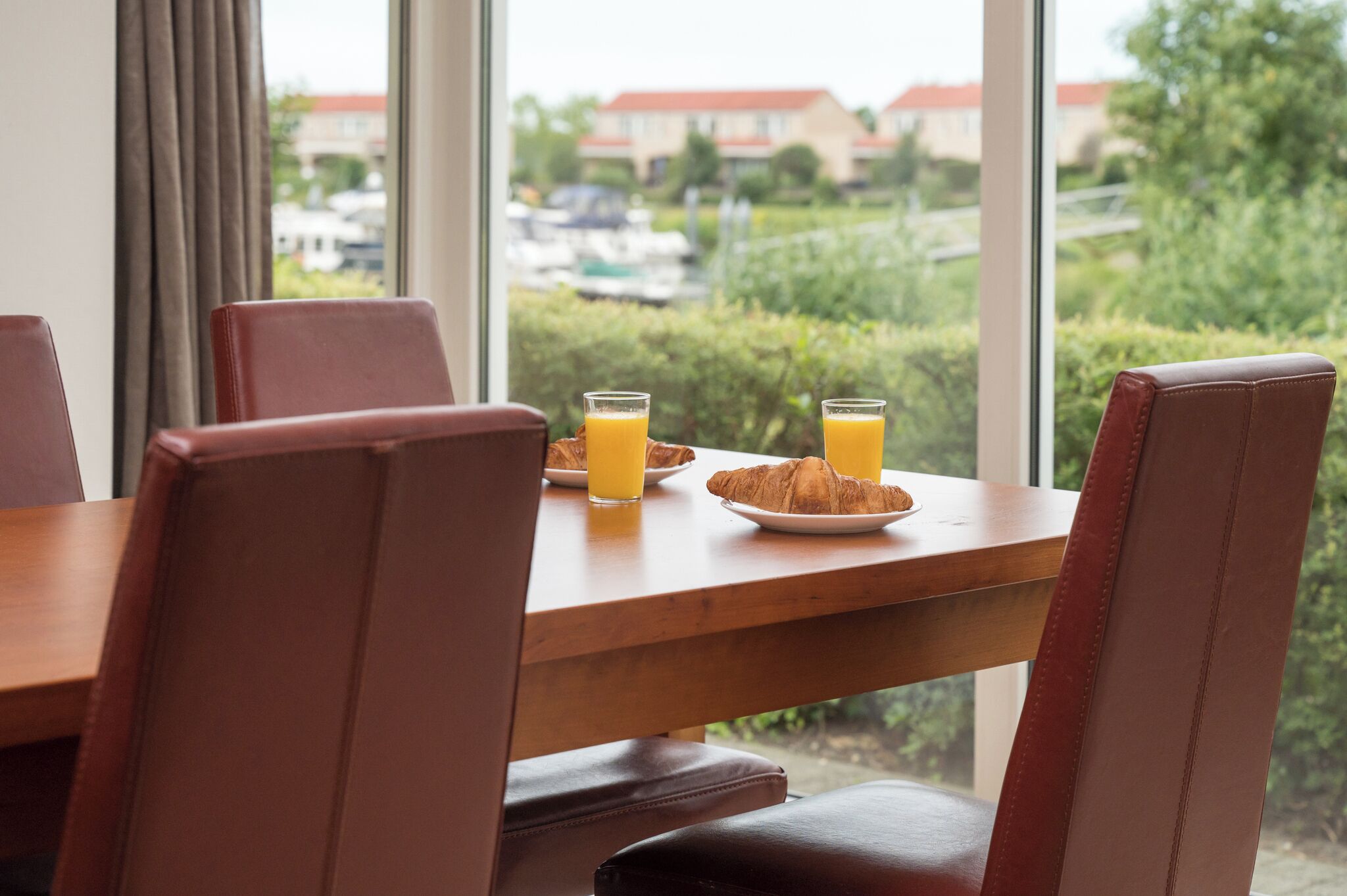 Una mesa con sillas, platos de cruasanes y jugo de naranja, y una vista de follaje y casas a través de grandes ventanales.